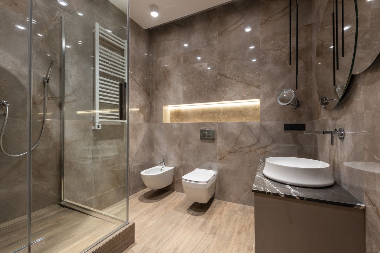 een badkamer met grijze tegels en beige vloer met toilet, bidet en douche. Ook duidelijk te zien is een nis in de muur die speciaal verlicht is
